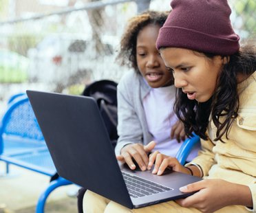 two girls in a playground looking at a laptop screen with shocked expressions on their face